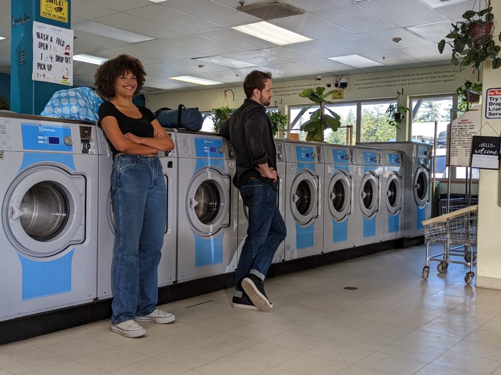 A young woman of color and a white guy stand at a bank of washing machines. She is smiling, he is turned away from her. 