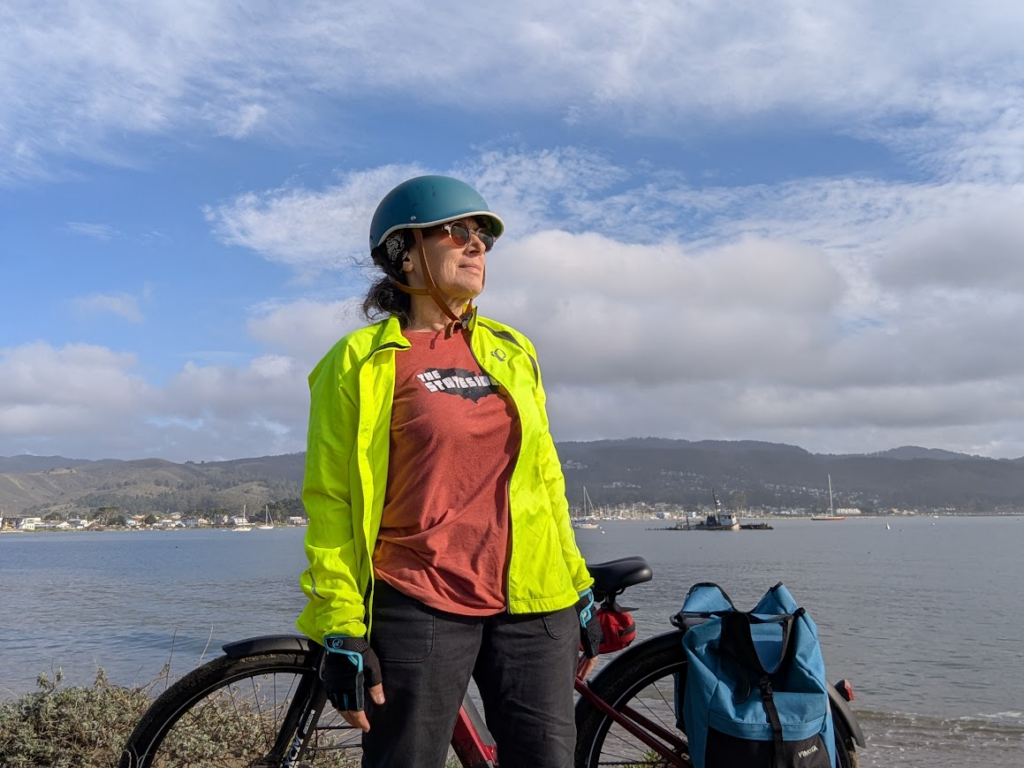 Pam in a bike helmet and safety yellow jacket in front of her bicycle on the shores of Half Moon Bay. 
